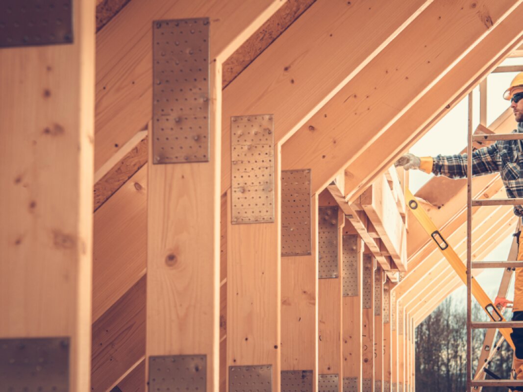 Un homme portant un casque de sécurité, des lunettes de protection, une chemise à carreaux et un pantalon de travail jaune est debout sur une échelle à l'intérieur de la **structure de toit massive et fortement charpentée en bois** d'une maison en construction. Il tient un niveau à bulle jaune et inspecte attentivement l'installation précise des composants en bois robustes qui forment l'impressionnante ossature du toit. La scène met l'accent sur la solidité et l'échelle de cette première étape de la construction d'une maison en bois.