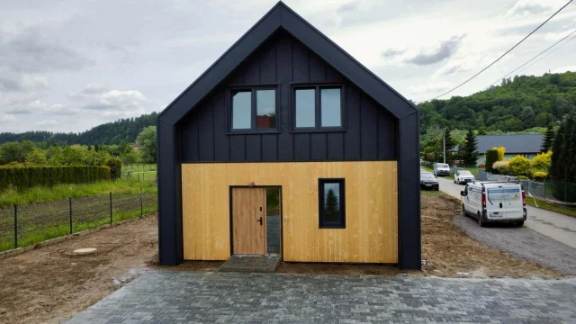 Front Ekodom110: czarny dach, jasna elewacja, zielone wzgórza.