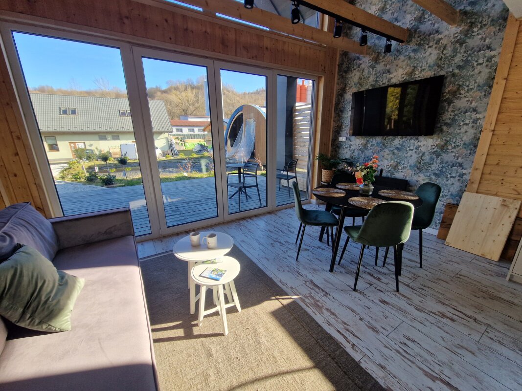 Salon z aneksem w Ekodom50: szara sofa, okrągłe stoliki, stół, TV, dekoracje.