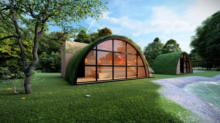 Moderne Glashaus-Sauna mit Gründach und panoramaartiger, abgerundeter Verglasung, harmonisch in die grüne und baumreiche Umgebung integriert, mit sichtbarem Interieur mit Holzbänken. Im Hintergrund befindet sich ein kleineres, ähnliches Objekt mit Gründach, und daneben führt ein Weg zu einem Wasserbecken.
