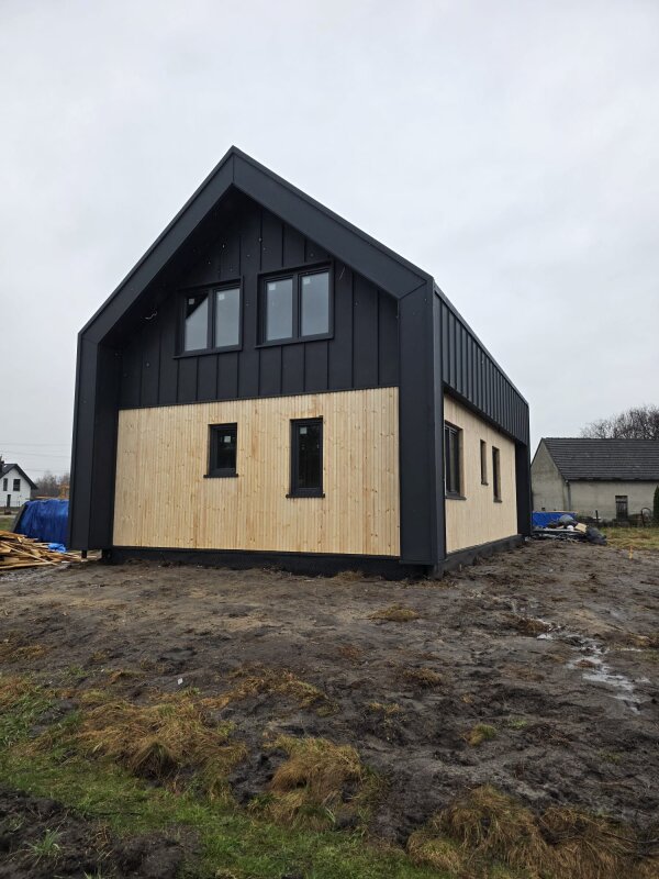 Dom szkieletowy w trakcie budowy z zewnątrz, widoczna elewacja z jasnej deski świerkowej i ciemne wykończenie blachą na rąbek