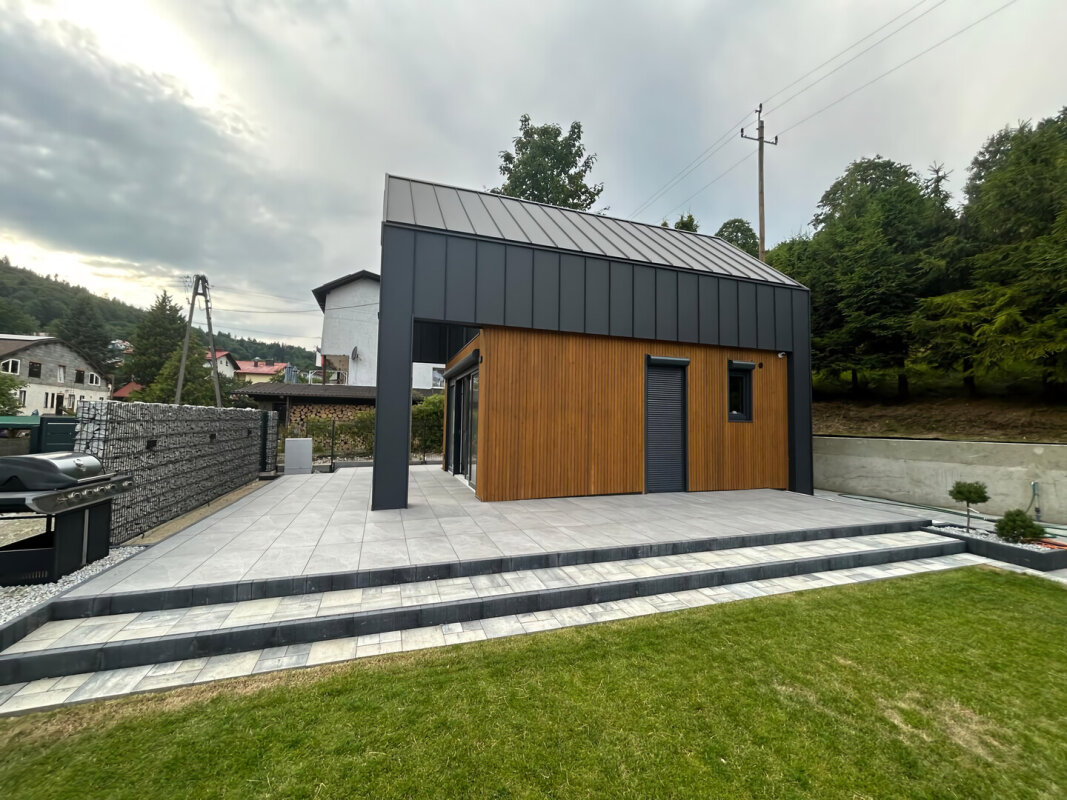 Widok boczny domu szkieletowego, taras z płyt gresowych i nowoczesne gabiony.