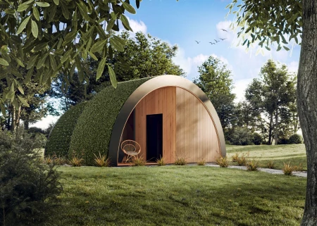 Modern Wellness arc sauna in a semi-circular shape, with a natural wood front and partially covered with green vegetation. A wicker armchair stands in front of the entrance, and the building harmoniously blends into the surroundings of a green lawn and trees under a blue sky.